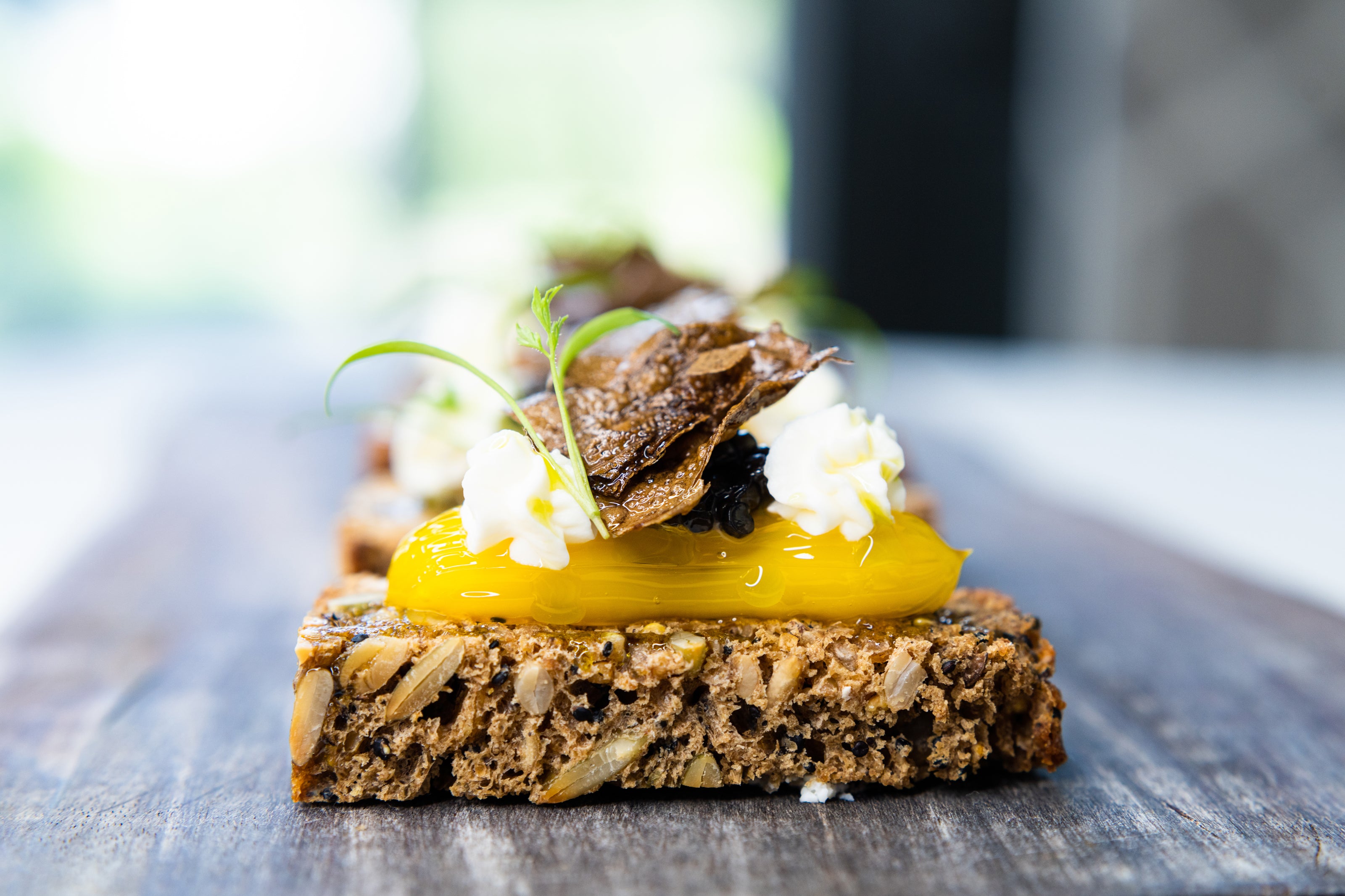 A close-up of a slice of multigrain bread topped with a bright yellow sauce, creamy spread, herbs, and a crispy garnish, placed on a wooden board with a blurred background.