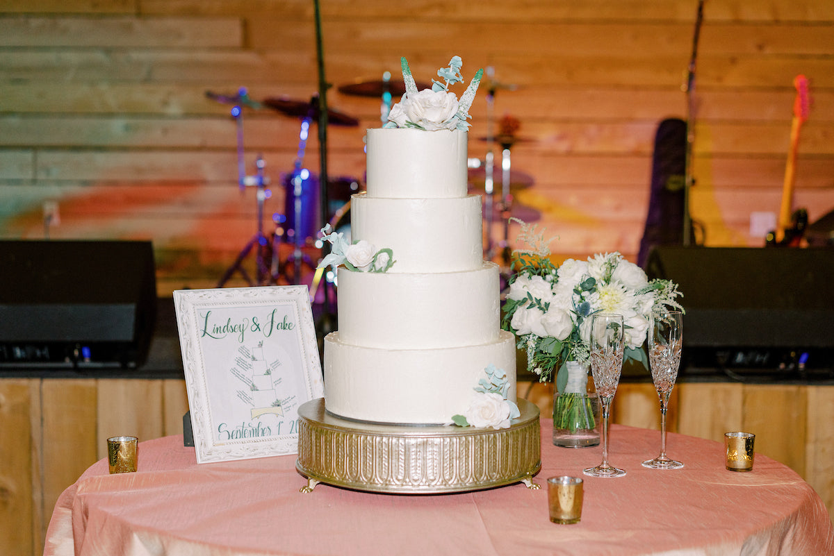 A four-tiered white wedding cake with floral decorations sits on a stand, surrounded by white flowers, two champagne flutes, a framed sign, and candles on a pink tablecloth, with a stage and instruments in the background.