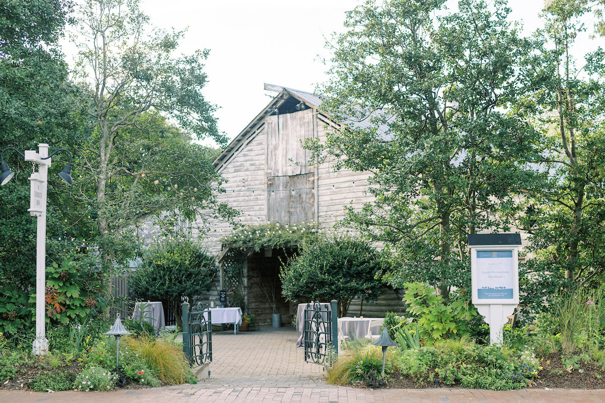 A rustic wooden barn surrounded by lush greenery and trees, with tables covered in white cloths set outside its entrance on a brick path. A sign is visible on the right, and lanterns and plants frame the scene.