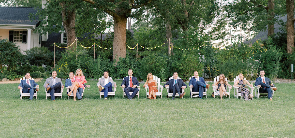 A group of people dressed in formal attire sit in a row of white chairs on a lawn, with large trees and string lights in the background, creating an outdoor, festive atmosphere.