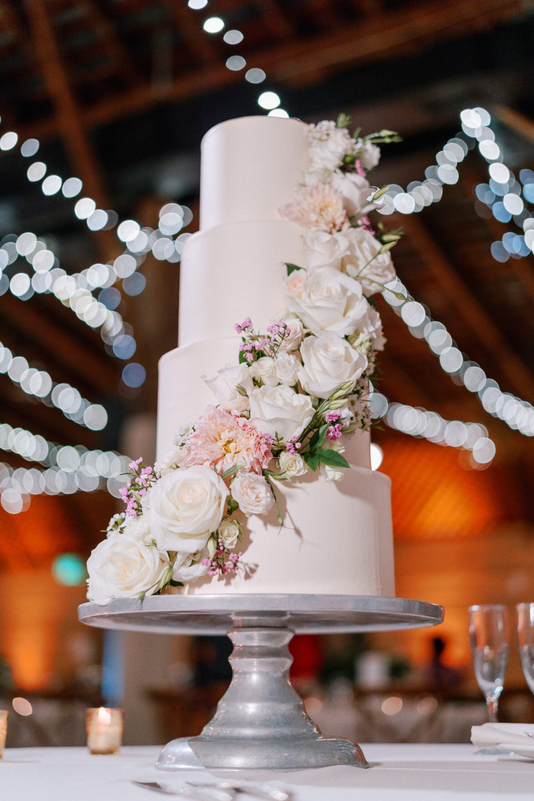 A four-tiered white wedding cake decorated with cascading white roses and pink flowers stands on a silver cake stand. String lights hang and glow softly in the blurred background.