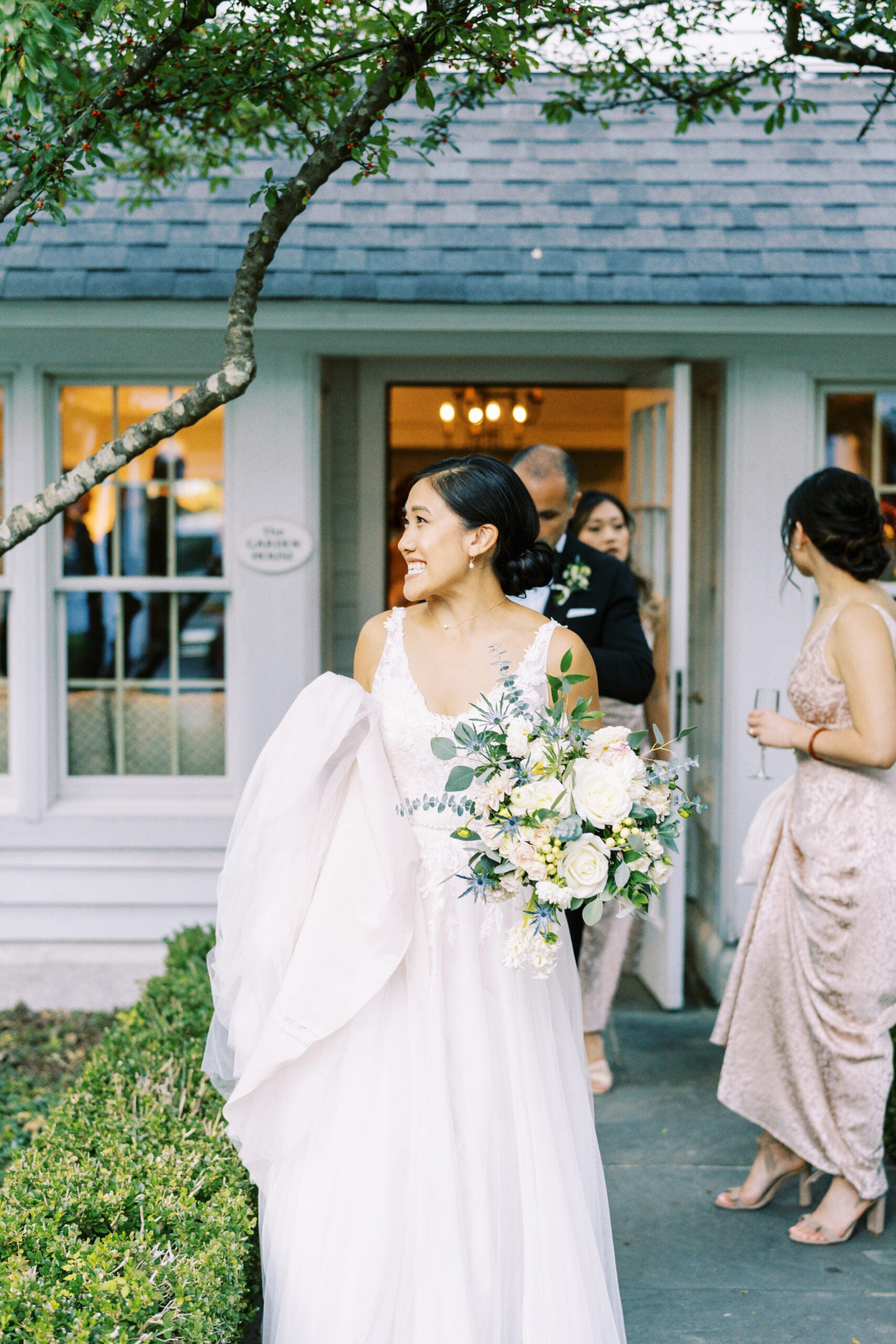A bride in a white dress holding a bouquet of white roses smiles and looks to the side outside a building, while guests in formal attire stand in the background.