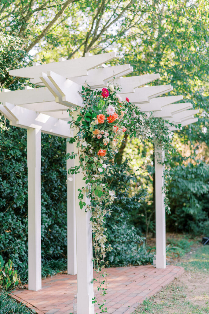 A white wooden pergola adorned with a lush floral arrangement of orange, pink, and white flowers with trailing greenery, set on a brick platform in a garden surrounded by trees and bushes.