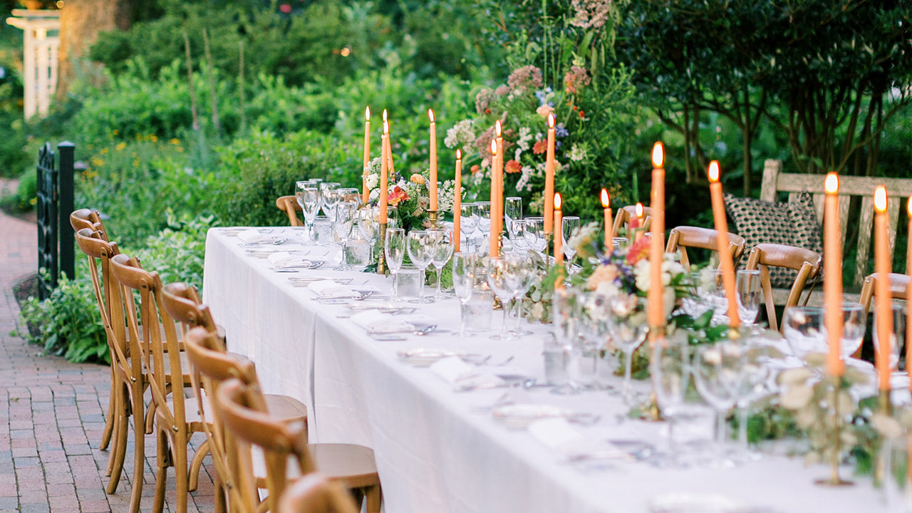 A long outdoor table set for a formal event is decorated with lit taper candles, glassware, floral centerpieces, and wooden chairs, surrounded by lush greenery and a garden setting.