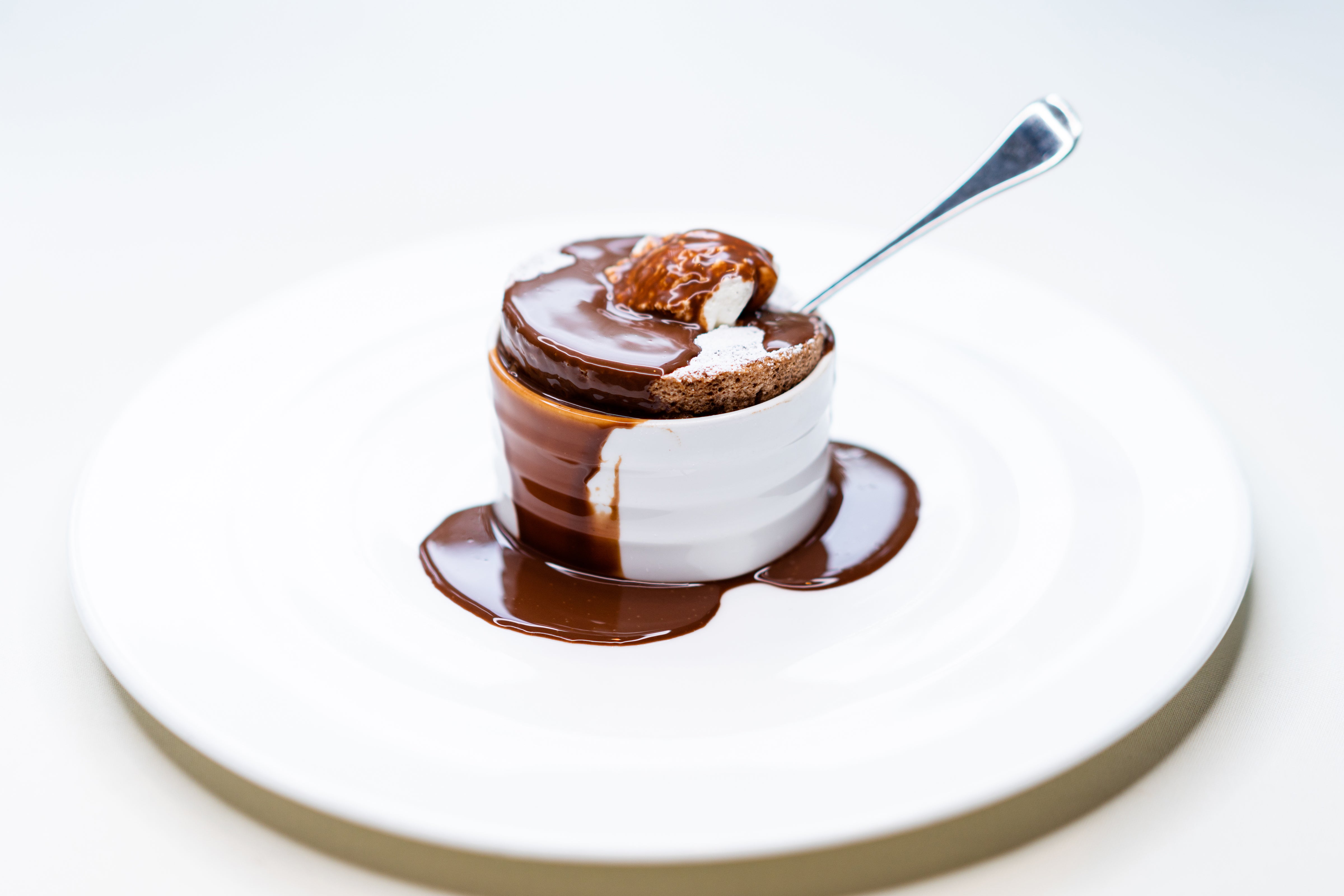 A chocolate soufflé in a white ramekin sits on a white plate, topped with a scoop of ice cream and drizzled with melted chocolate, with a spoon inserted into the dessert.