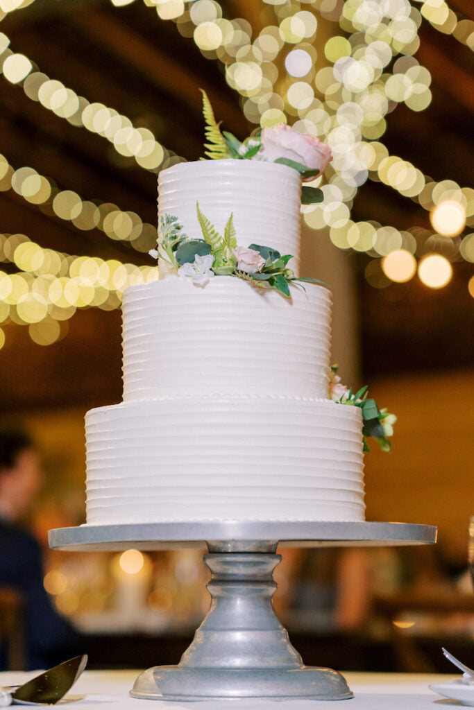 A three-tiered white wedding cake decorated with green ferns and pink flowers sits on a silver cake stand, with blurred string lights creating a warm, festive background.