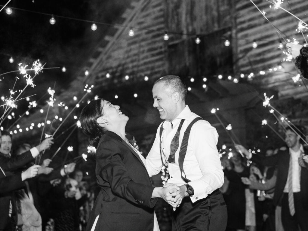 A joyful couple dances at night under string lights, surrounded by guests holding sparklers. The scene is festive and lively in front of a rustic barn. Both are smiling and laughing, dressed in formal attire.