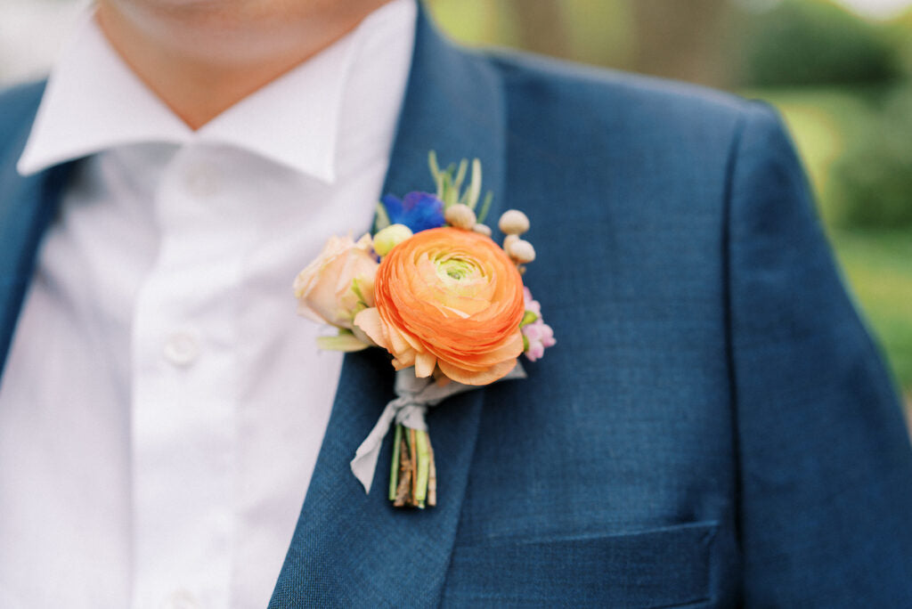 A close-up of a person in a blue suit and white shirt, wearing a boutonnière with an orange ranunculus flower and small accent blossoms pinned to their lapel. The background is blurred greenery.