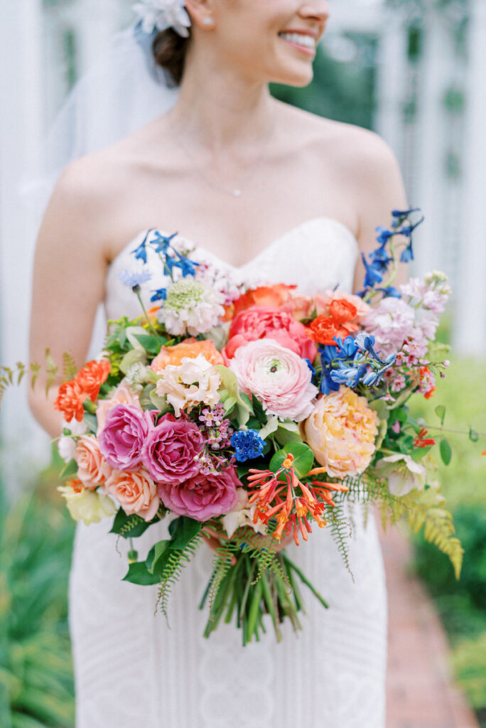 A bride in a strapless white dress holds a vibrant bouquet of colorful flowers, including roses, peonies, and greenery, while smiling outdoors. The background is softly blurred.