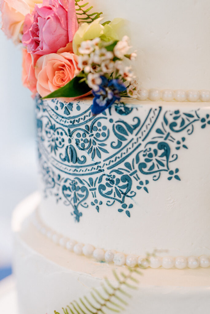 Close-up of a white tiered cake decorated with blue intricate floral patterns, pearl-like icing borders, and topped with pink, peach, and white flowers, as well as a green fern leaf.