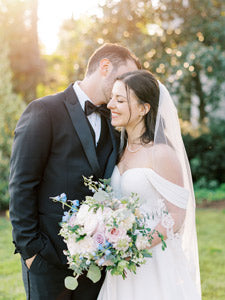 A groom in a black tuxedo kisses the bride’s forehead as she smiles, holding a pastel floral bouquet. The bride wears an off-the-shoulder white gown and veil. They stand outdoors with sunlight and greenery in the background.