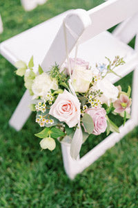 A small bouquet of pastel flowers, including roses and greenery, hangs from a white ribbon on the back of a white folding chair set on green grass.