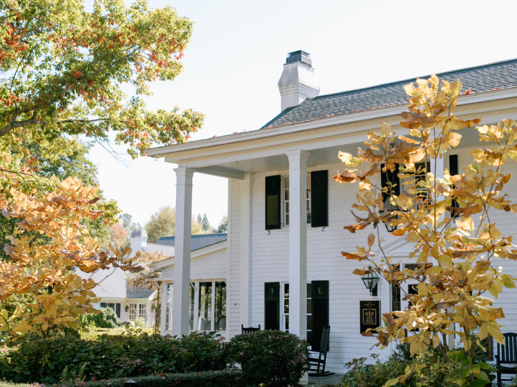 A white house with tall columns and black shutters is surrounded by autumn trees with yellow leaves. Two black rocking chairs sit on the front porch in the sunlight.
