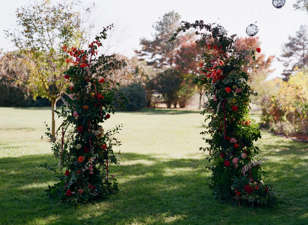 Two tall, curved floral arrangements with greenery and vibrant red and orange flowers stand on green grass in a sunlit outdoor setting, with trees and shrubs in the background.