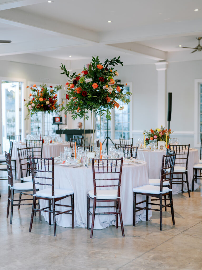 Elegant event space with round tables covered in white tablecloths, surrounded by dark wooden chairs. Tall floral centerpieces with vibrant red, orange, and yellow flowers decorate each table. The room is bright and spacious.