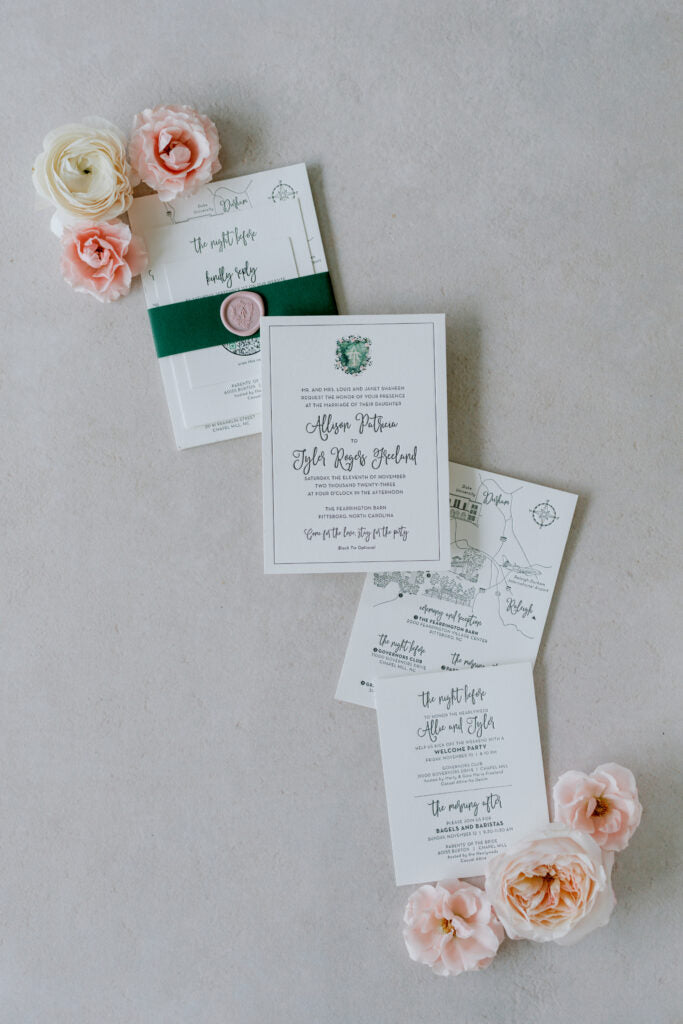 Flat lay of elegant wedding invitations with floral designs, surrounded by blush pink and cream roses on a light grey surface. The invitations are neatly stacked, with some overlapping and a green wax-sealed band.