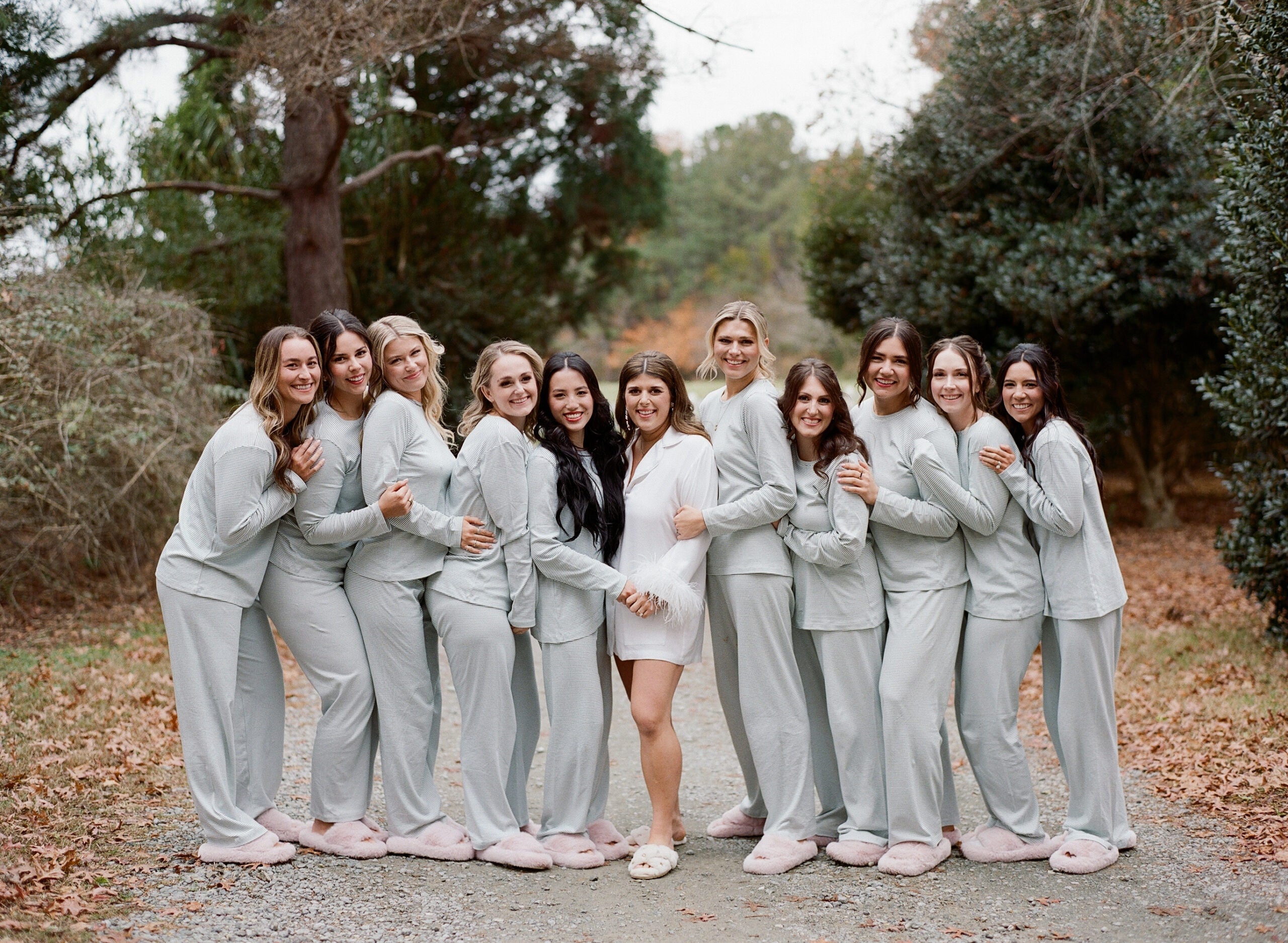 A group of eleven women stand outdoors on a path surrounded by trees. Ten wear matching light gray pajamas and pink slippers, while the woman in the center wears a white outfit and slippers. They are smiling and posing together.