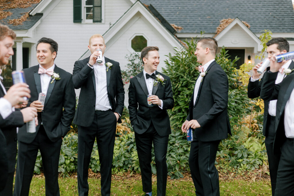 A group of men in black tuxedos stand outside in front of a house, smiling and talking while holding drinks, likely before a formal event or wedding.