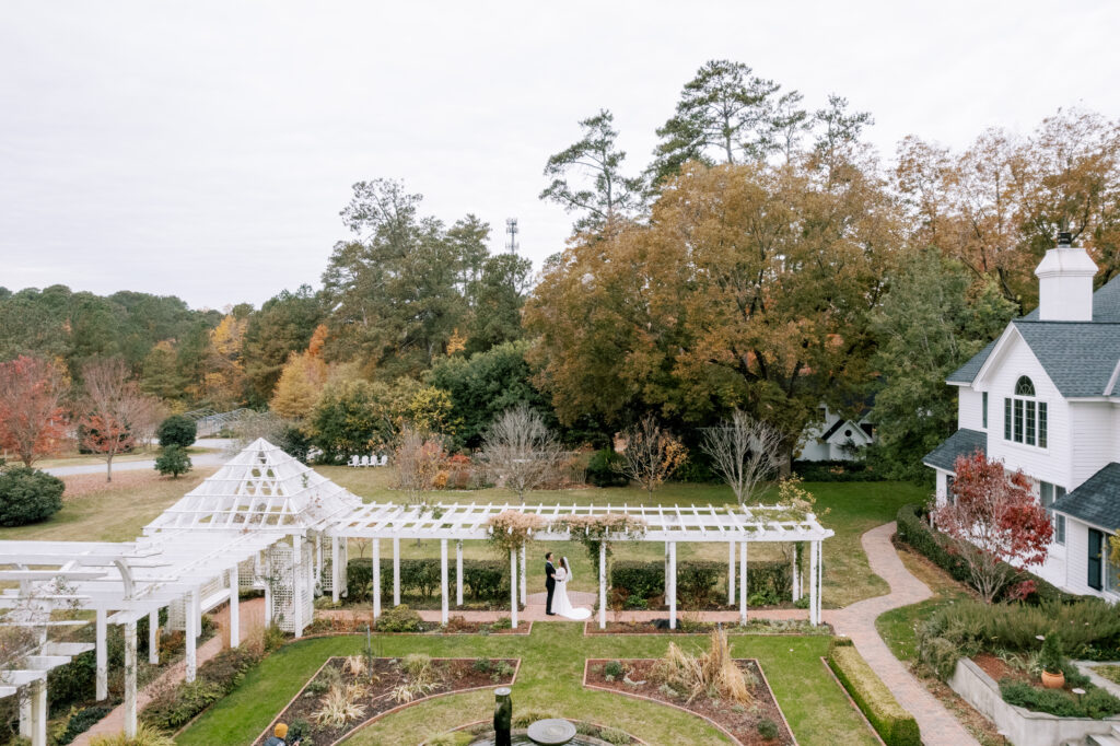 A bride and groom stand under a white pergola in a garden, surrounded by autumn trees, greenery, and a white house with a dark roof on the right. The scene is calm and picturesque.