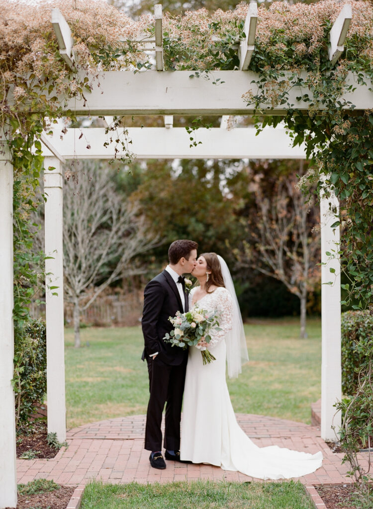 A bride and groom share a kiss under a white pergola covered in greenery and flowers in a garden. The bride wears a white gown and veil, holding a bouquet, while the groom wears a black suit and tie.