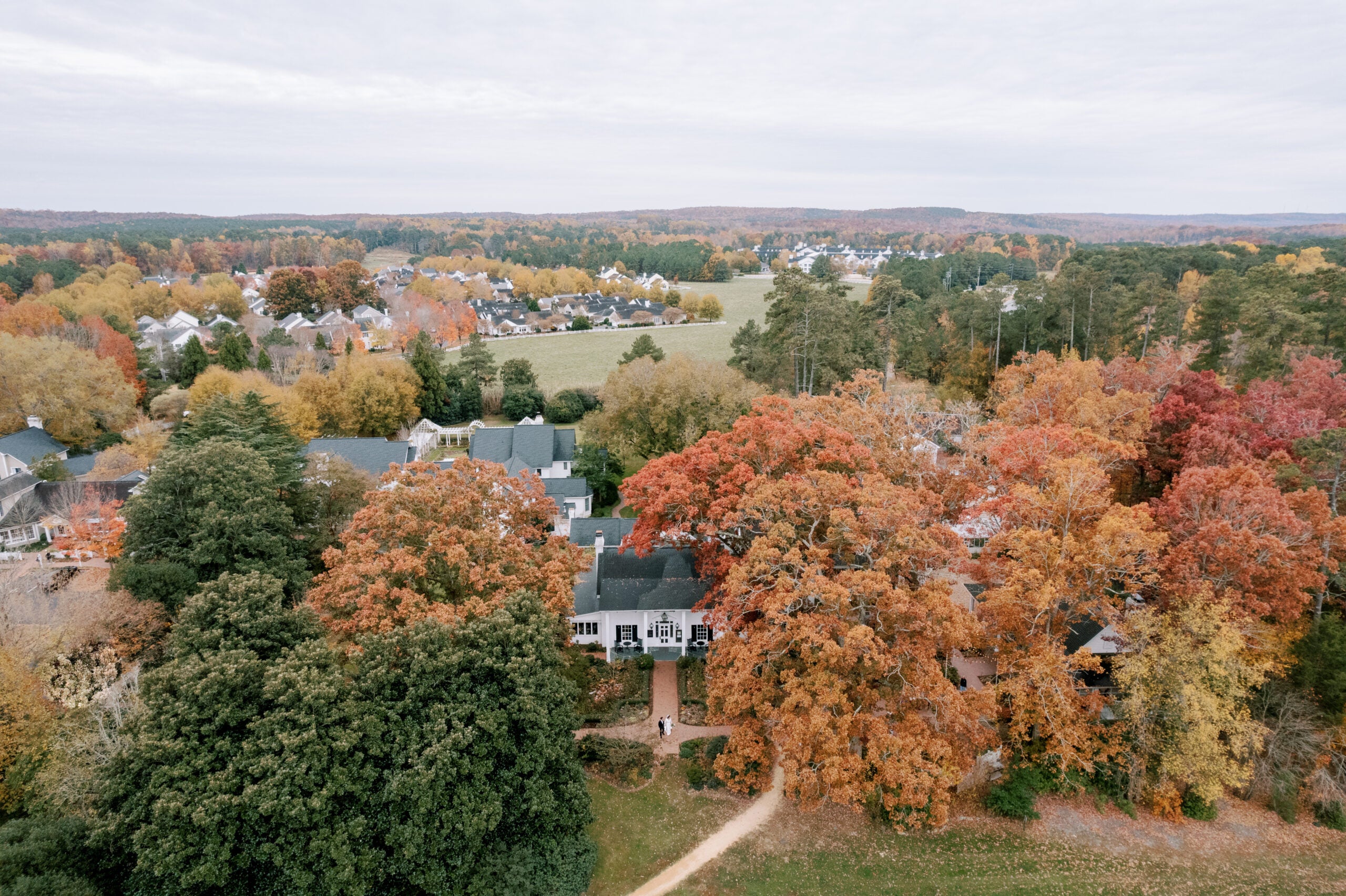 Aerial view of a suburban neighborhood surrounded by autumn trees with colorful foliage in shades of red, orange, and yellow, and open green fields in the distance under a cloudy sky.