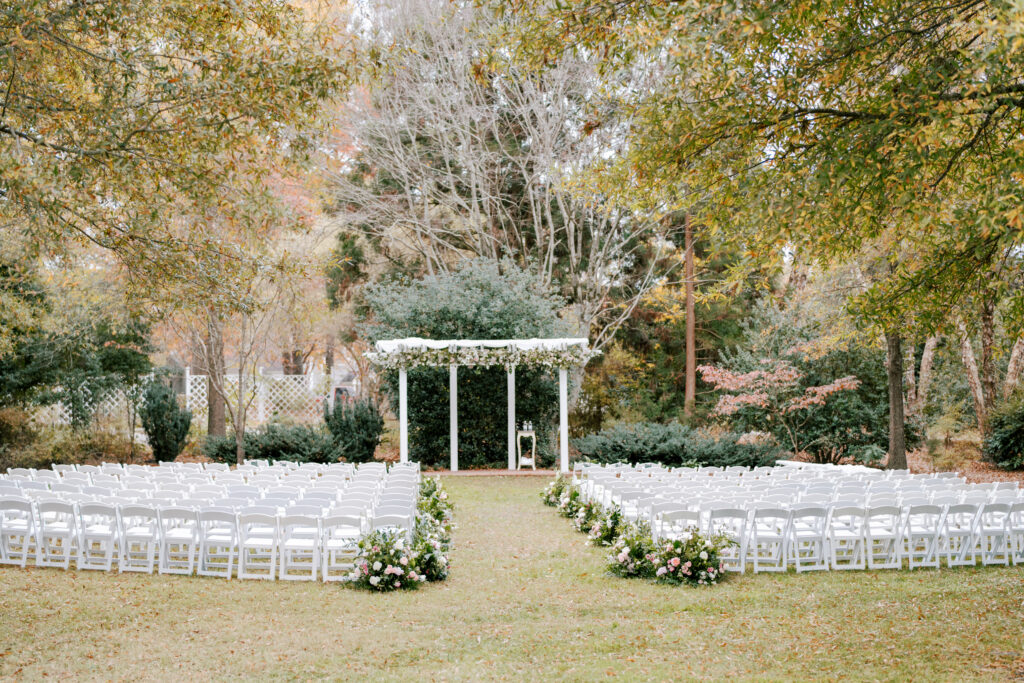 An outdoor wedding setup with rows of white chairs facing a white pergola adorned with greenery and flowers, surrounded by trees and flowers on a grassy lawn.