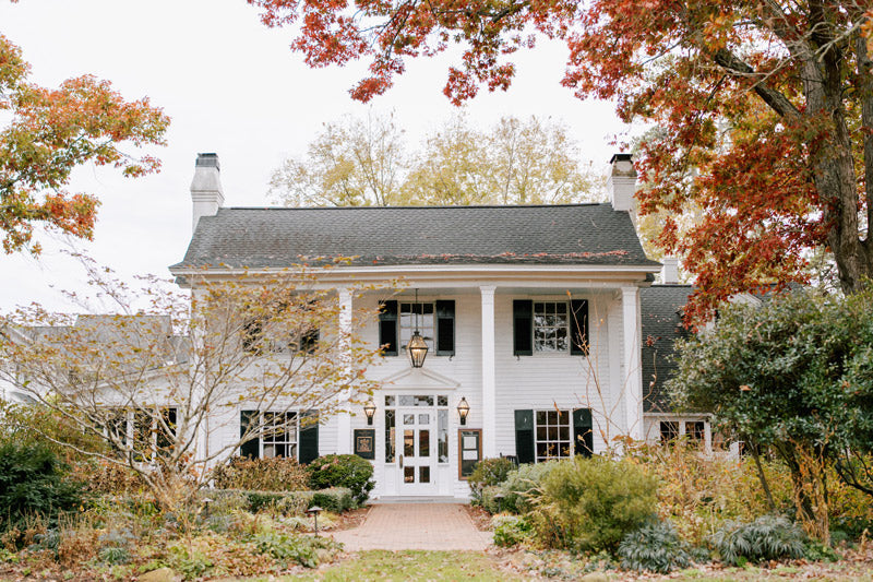 A large white colonial-style house with black shutters and two chimneys, surrounded by autumn trees and greenery, viewed from the brick path leading to the front door.