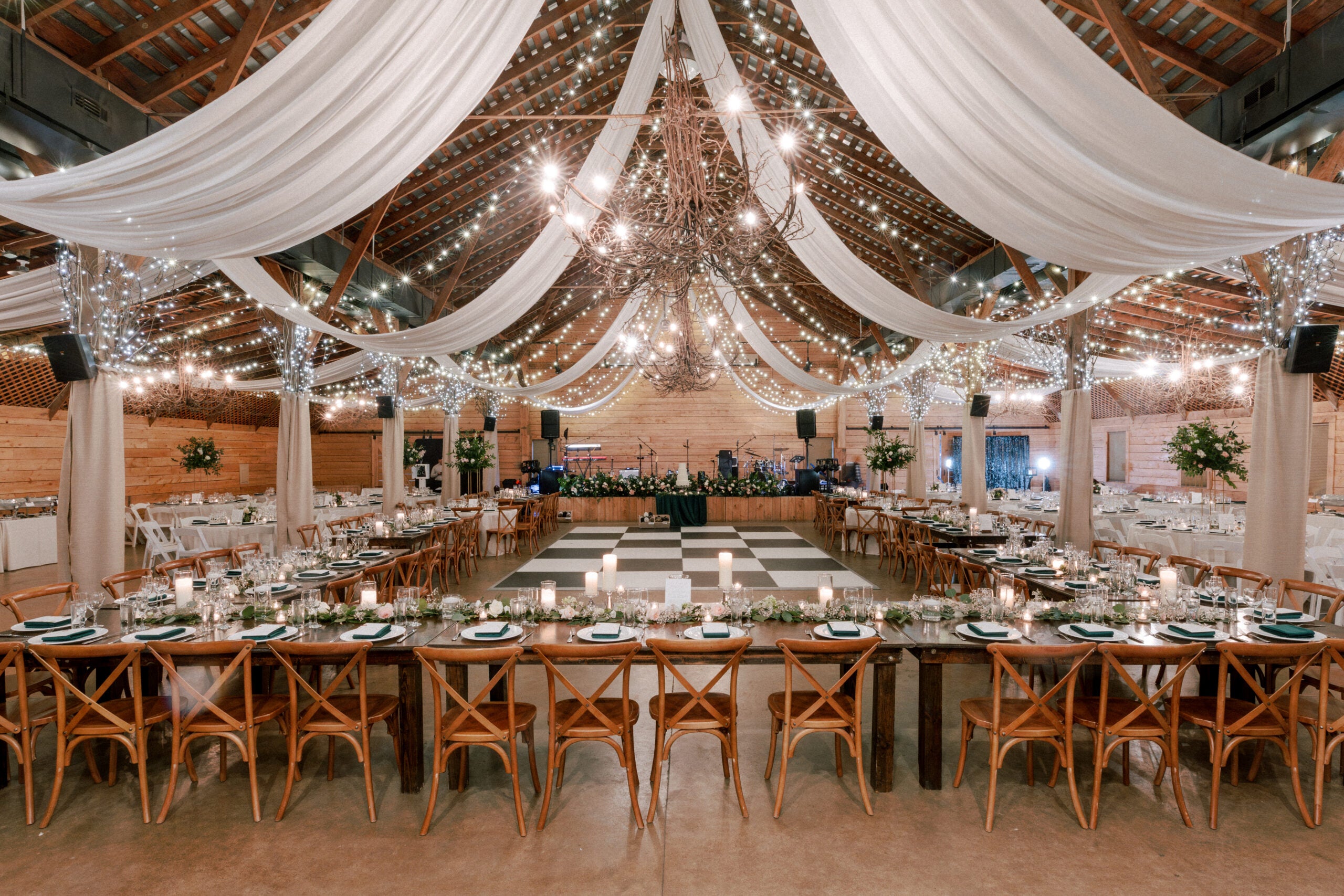 A rustic wedding venue is decorated with draped white fabric, string lights, and chandeliers. Long wooden tables with elegant place settings form a U-shape around a checkered dance floor.