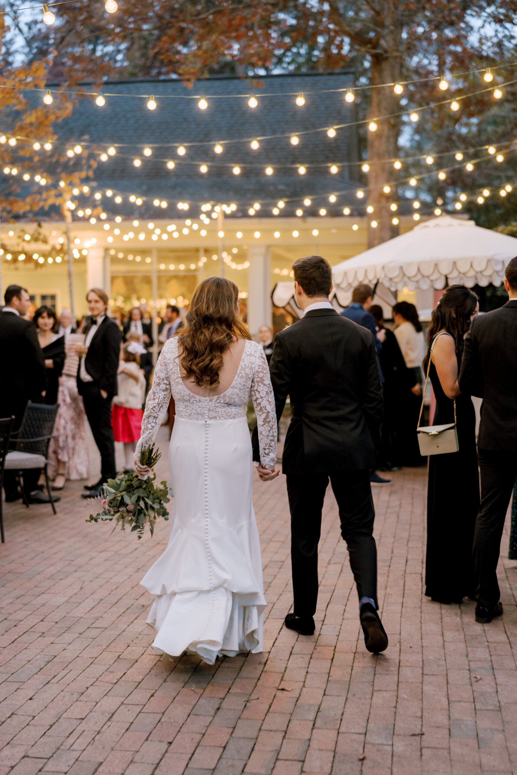 A bride in a white dress holding a bouquet and a groom in a black suit walk hand in hand at an outdoor wedding reception with string lights and guests socializing in the background.