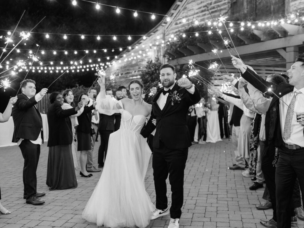 A bride and groom walk hand in hand under string lights, smiling as guests hold sparklers and celebrate around them during an outdoor evening wedding.