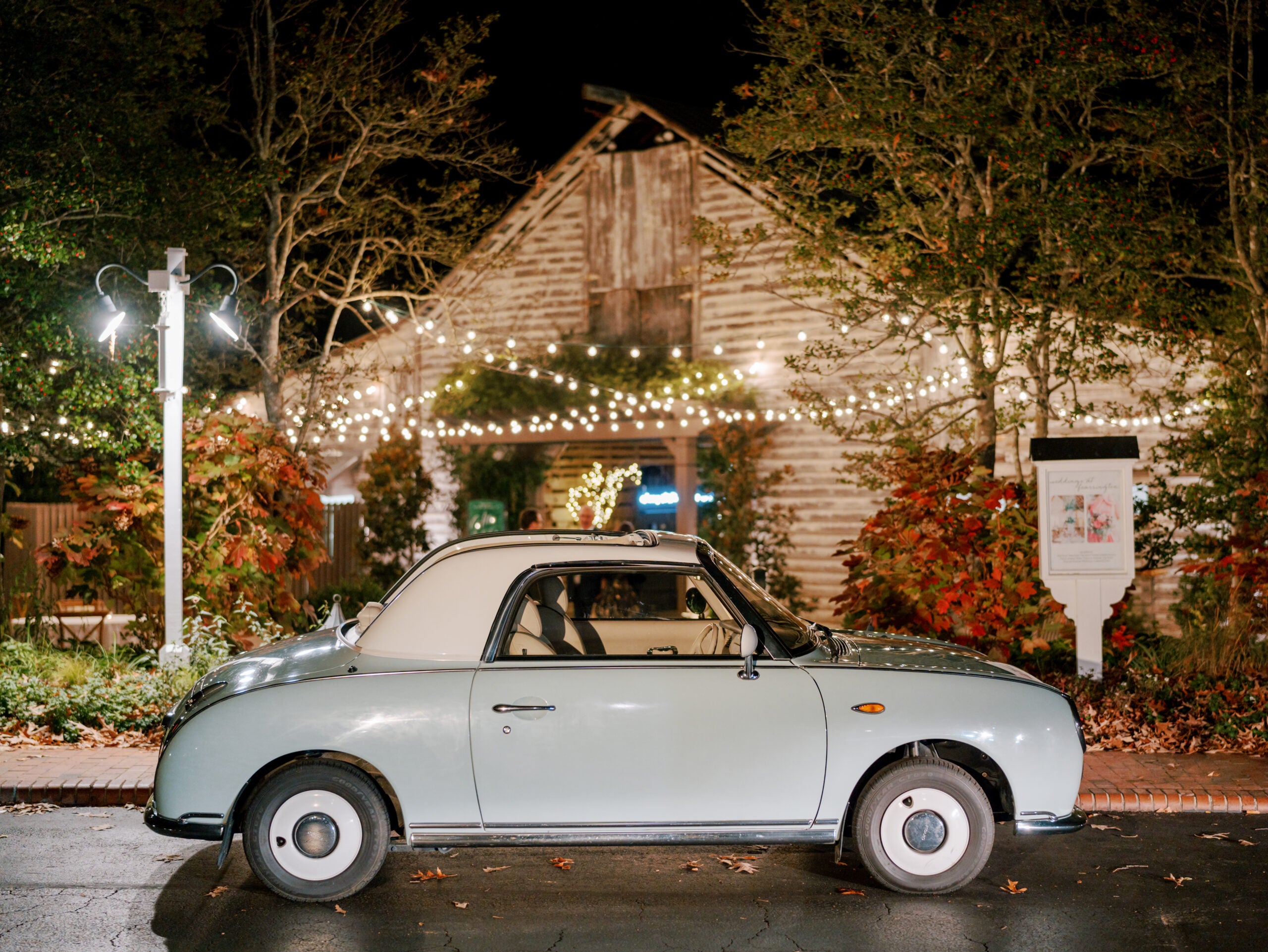 A vintage light blue car is parked on a wet street at night in front of a rustic wooden building with string lights and autumn foliage.