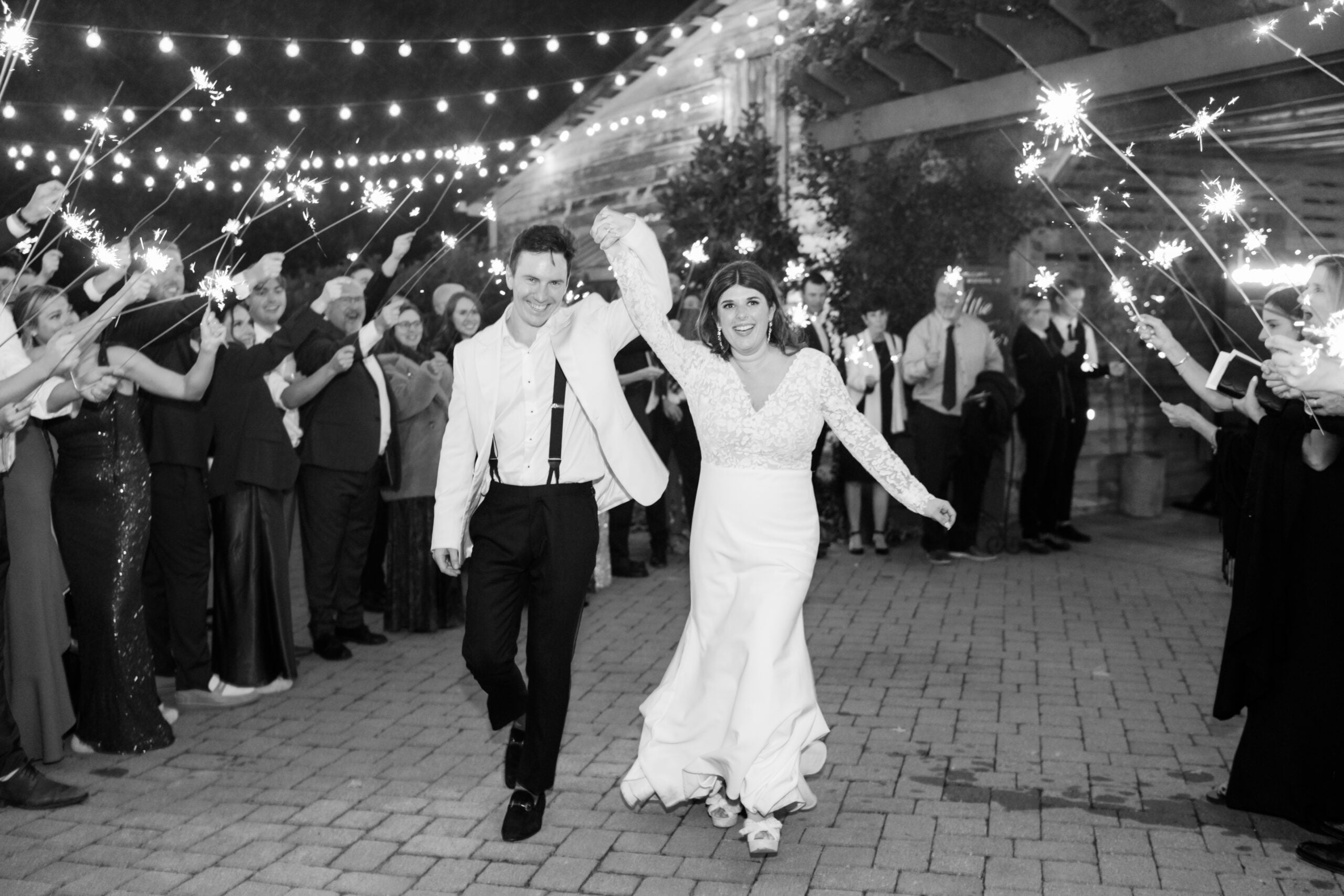 A newlywed couple, smiling and holding hands, walks under string lights as guests cheer and wave sparklers during an outdoor nighttime celebration.