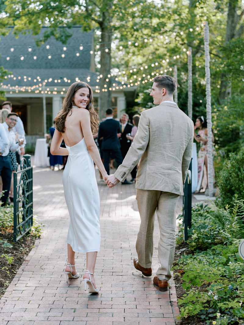 A couple holding hands walks down a garden path at an outdoor event, with the woman in a white dress smiling back at the camera. String lights and guests are visible in the background.