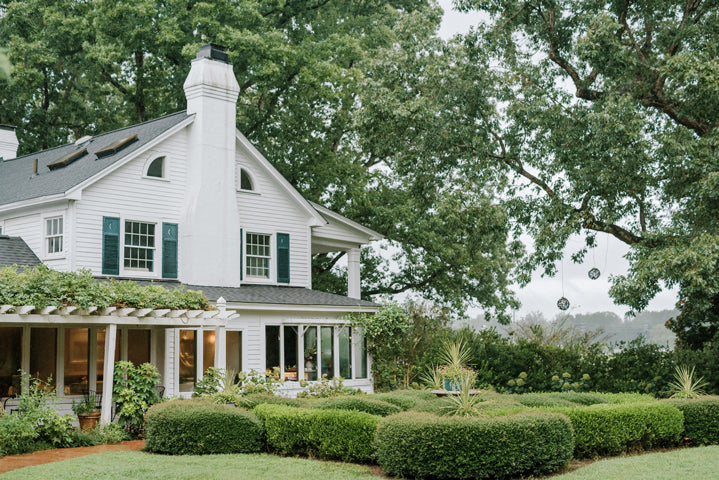 A white two-story house with dark shutters sits surrounded by lush green bushes and trees. Large windows and a pergola cover a patio area, while trimmed hedges add to the well-kept garden.