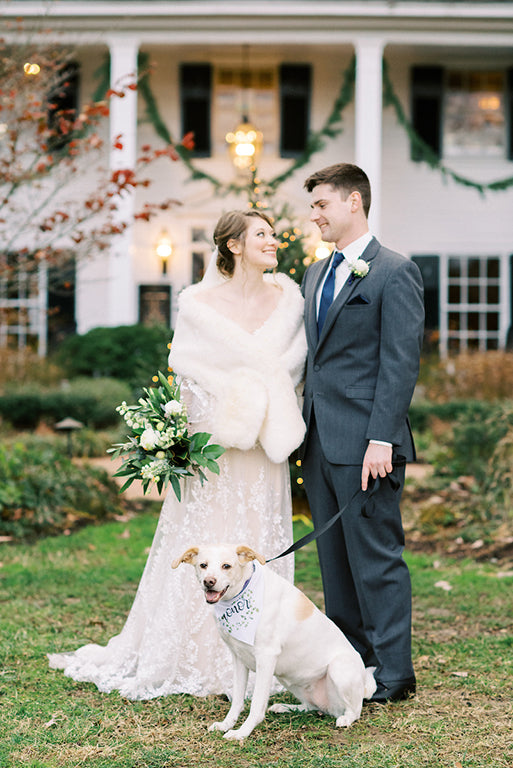 A bride and groom stand smiling at each other outside, with greenery and a large white house behind them. The bride holds a bouquet and wears a white dress; the groom wears a suit. A dog with a 