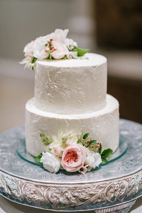 Two-tiered white wedding cake with textured frosting, decorated with pink and white flowers on top and at the base, displayed on an ornate silver cake stand.