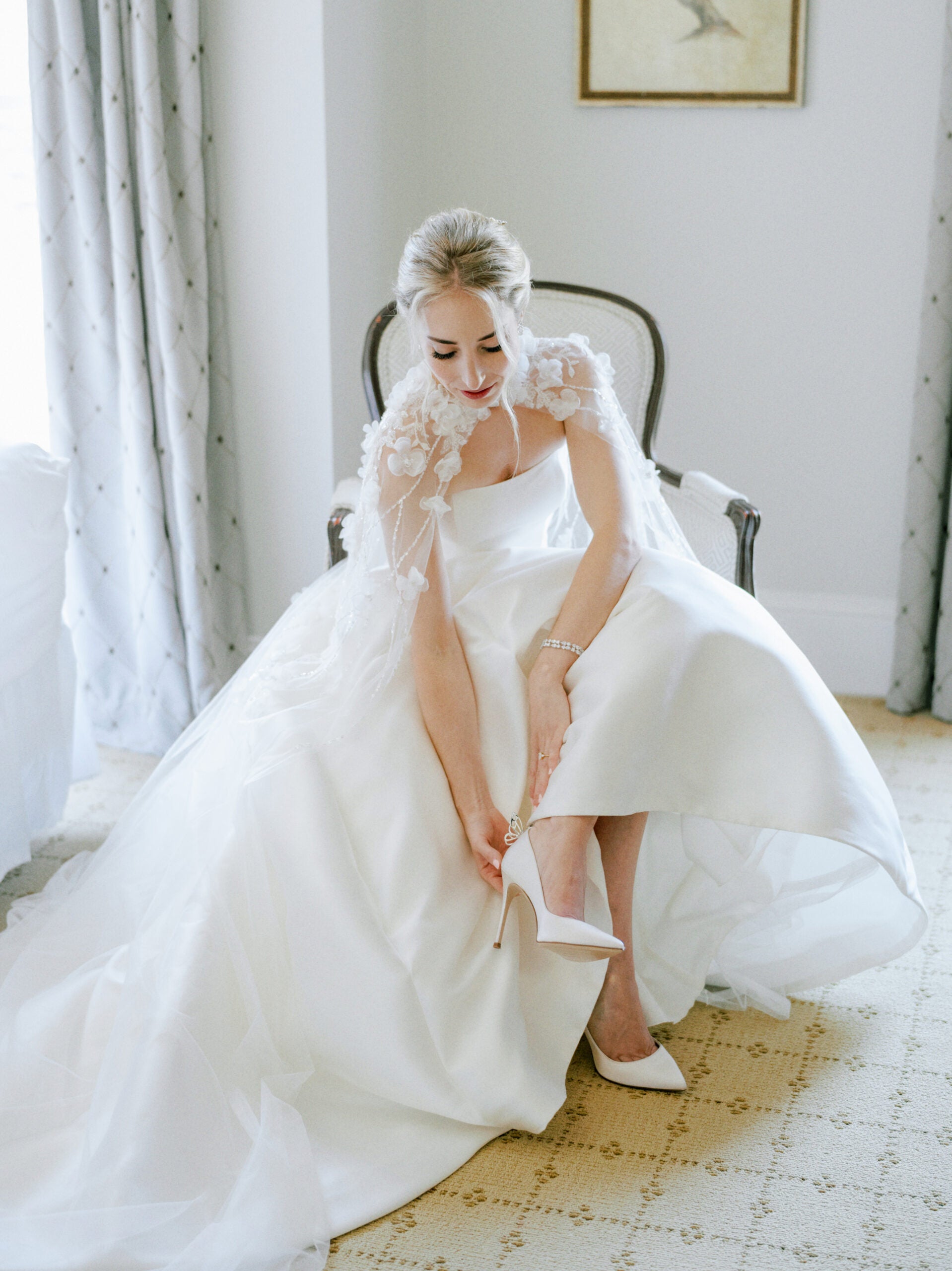 A bride in a white wedding gown sits on a chair, looking down as she fastens the strap on her white high-heeled shoe. Soft light fills the elegant room, and a delicate floral veil drapes over her shoulders.