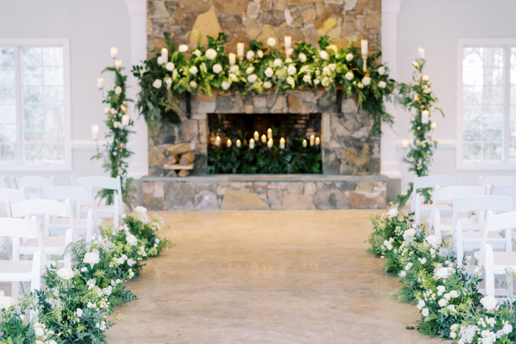 A wedding ceremony setup with white chairs arranged in rows facing a stone fireplace, decorated with lush greenery, white flowers, and candles on the mantle and along the aisle.