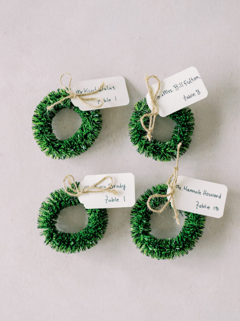 Four miniature green wreaths with twine bows, each holding a white tag with handwritten names and table numbers, are arranged on a light surface.