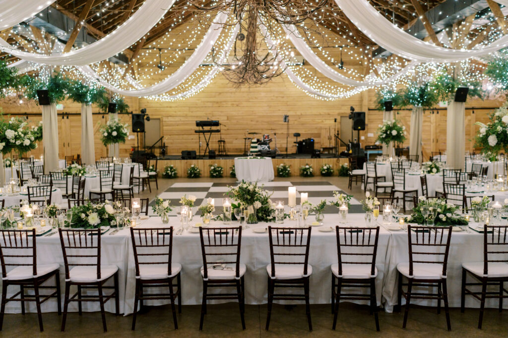 Elegant wedding reception hall with long tables set for dinner, decorated with white flowers and greenery. String lights and draped fabric hang from the wooden ceiling above a dance floor and stage.