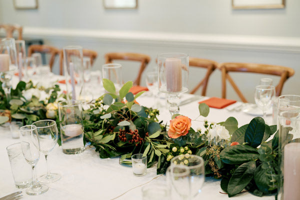 A close-up of a table set for an event, featuring a white tablecloth, glassware, candles, orange napkins, and a floral centerpiece with greenery and orange and white flowers. Wooden chairs line the table.