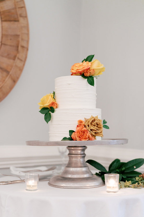 A two-tiered white cake with textured icing, decorated with orange and yellow roses, sits on a silver cake stand. The table is covered with a white cloth and small lit candles.