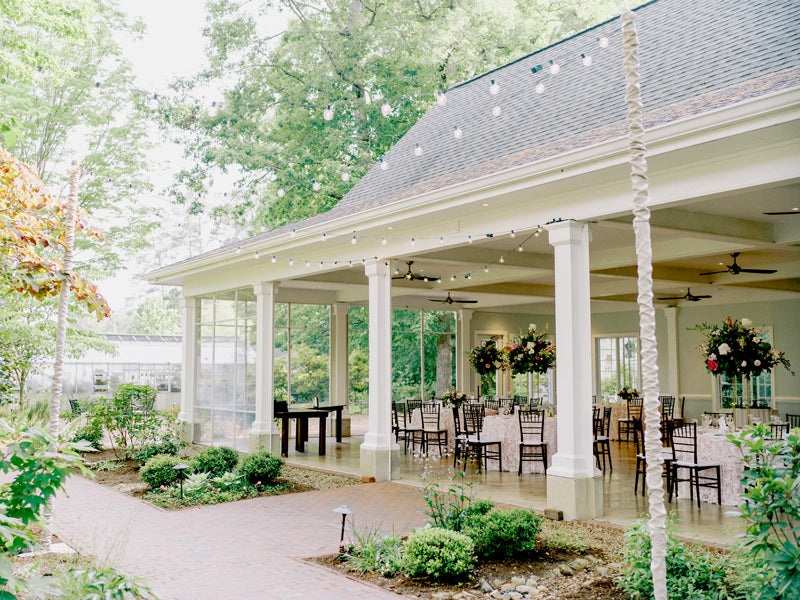 An elegant outdoor pavilion with string lights, surrounded by lush greenery. Inside, round tables with floral centerpieces and dark chairs are arranged for an event, and large windows let in natural light.