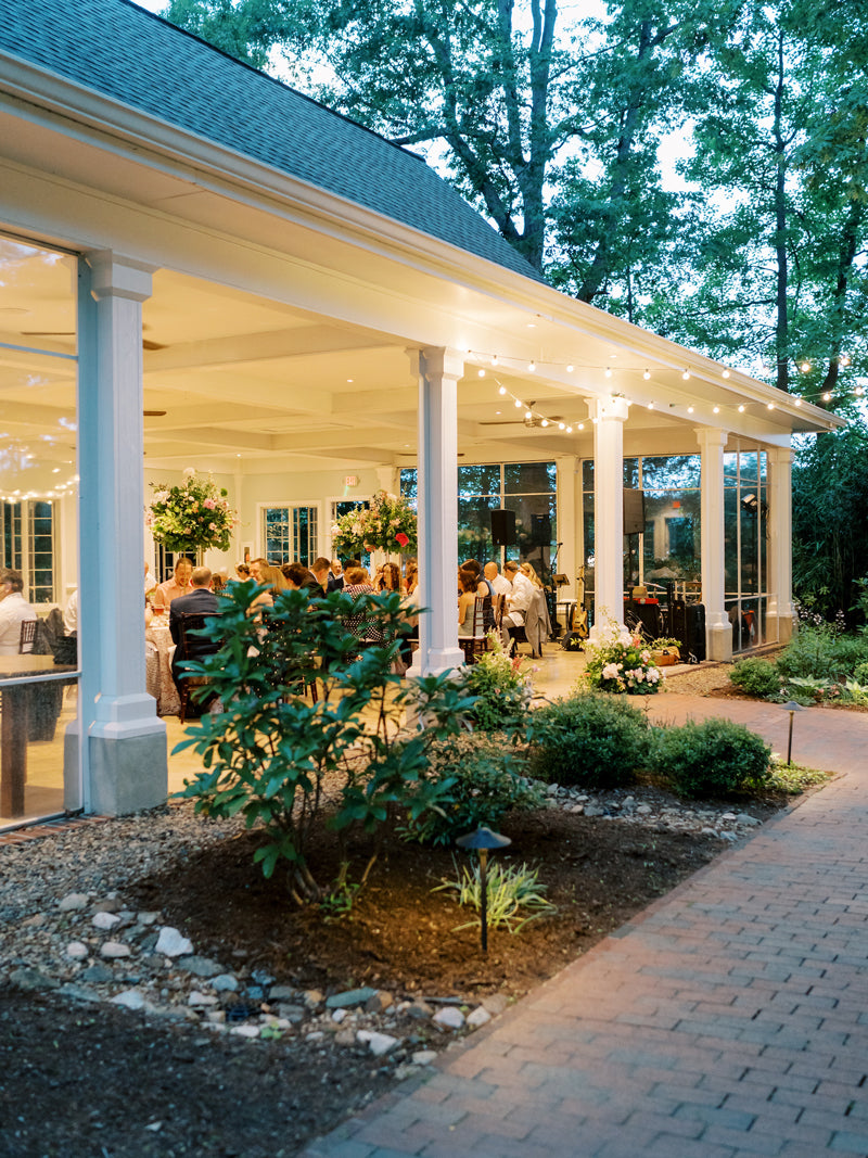 An elegant outdoor pavilion decorated with string lights and flowers, hosting a gathering of people dressed formally. Lush greenery and a brick path surround the venue as evening approaches.