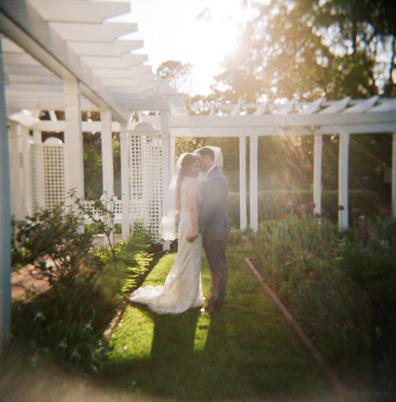 A bride and groom stand close together under a white pergola in a garden, with sunlight streaming through the structure, creating a soft and dreamy atmosphere.
