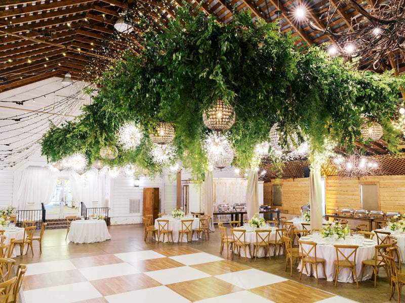 A spacious wedding venue with round tables and wooden chairs, decorated with white flowers. Lush greenery and sparkling lights hang from the ceiling, creating a warm, elegant atmosphere with a checkerboard dance floor beneath.