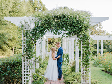 A bride and groom stand facing each other under a white, vine-covered arbor in a lush garden, surrounded by greenery and flowers, on a sunny day.