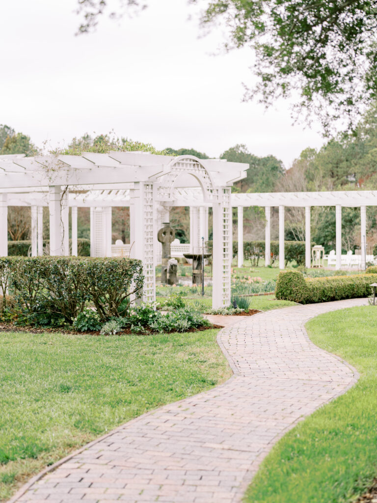 A winding brick path leads through a garden with neatly trimmed bushes and a white pergola structure, surrounded by green grass and trees in the background.