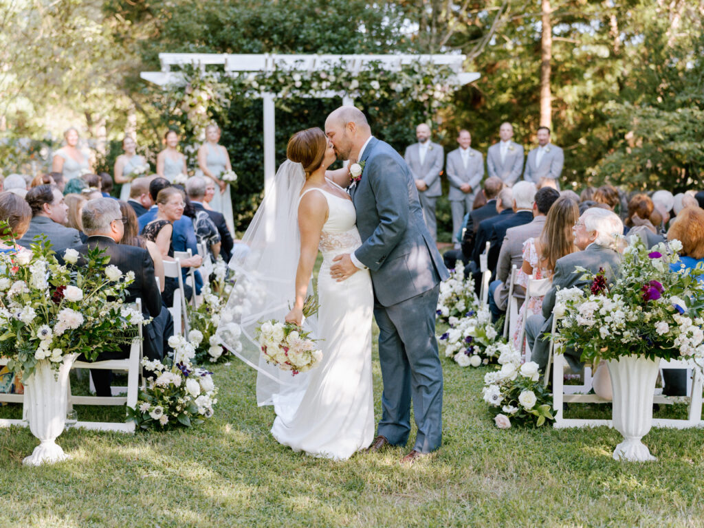A bride and groom share a kiss in the aisle during an outdoor wedding ceremony, surrounded by guests seated on either side and lush floral arrangements in a garden setting. Bridesmaids and groomsmen stand by the altar.