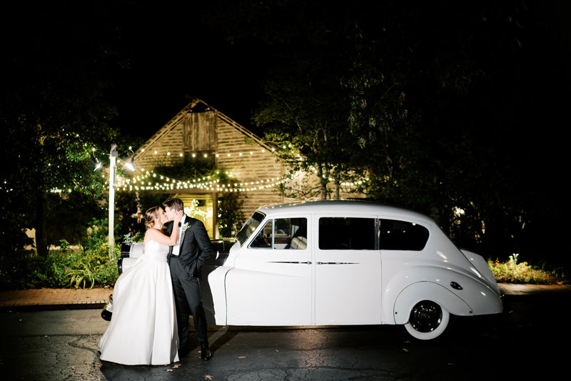 A bride and groom share a kiss at night beside a classic white car, with string lights and a rustic wooden building glowing softly in the background.